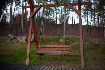 Large wooden garden swing bench on chains in a pine forest park with rocks
