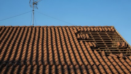 Red tiled roof with missing tiles and television antenna against clear blue sky terracotta clay