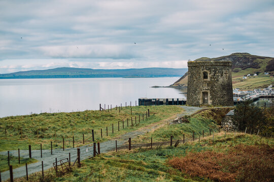 A historic landscape photograph featuring the circular Uig Tower, also known as Captain Fraser's Folly, perched on a hillside. The background reveals a sweeping view of Uig Bay (Loch Snizort) and the 