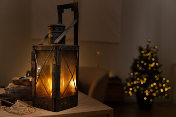 A vintage lantern casts a warm glow on a table next to a small Christmas tree.