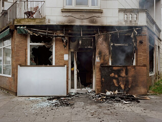 documentary-style photograph of a burned fruit shop storefront after a fire. The damaged brick facade shows shattered windows, charred walls, smoke stains, and debris scattered across the sidewalk.