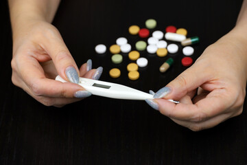 Woman measuring body temperature, concept of fevers symptoms. Digital thermometer in female hands and pills on  table