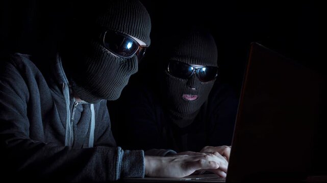 two robbers wearing balaclavas and sunglasses are working on laptops in a dark room
