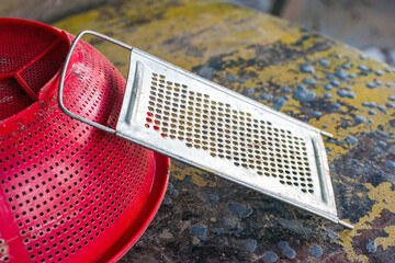 image features a close-up shot of a red colander and a metal grater, resting on a distressed and worn yellow surface, capturing a textural contrast of the everyday.