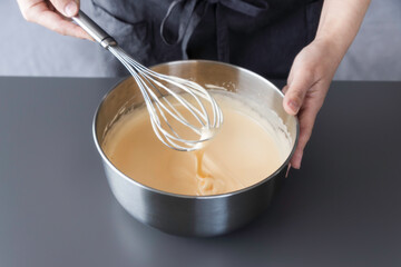 Hands whisking cake batter in a stainless steel bowl, batter dripping from whisk