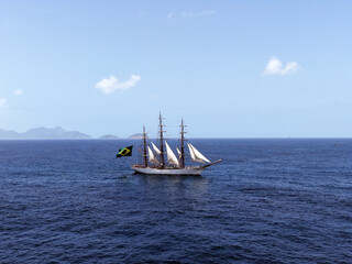 Aerial view of a three-masted sailing ship with sails and a large Brazilian flag on the stern sailing in the Atlantic Ocean. The training ship of the Brazilian Navy.