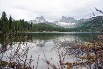 Calm mountain lake reflecting snow-capped peaks and green forest under a cloudy sky. Serene natural landscape for travel and nature concept.
