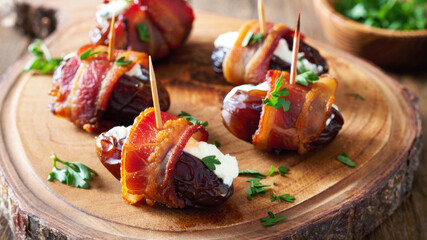 Bacon wrapped stuffed dates filled with soft cheese and fresh herbs, served as savory gourmet appetizers on a rustic wooden board, combining sweet fruit, crispy bacon, creamy texture for party food