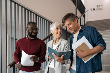 Three Professionals Discuss Documents In A Modern Office Hallway During Friendly Collaboration