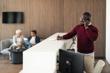 Busy Receptionist At Modern Office Front Desk Taking Notes While On A Phone Call