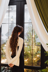 A girl with long wavy hair, wearing a white blouse and corset, stands and admires the view from the window.