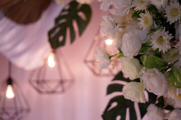 Elegant white artificial roses with geometric pendant lights and monstera leaves. Romantic floral decor for weddings, events, and modern aesthetic interiors