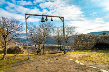 Fototapeta premium A simple metal frame holding three church bells stands among bare trees and stone ruins near Jvari Monastery, overlooking rolling hills under a bright blue sky with soft clouds.