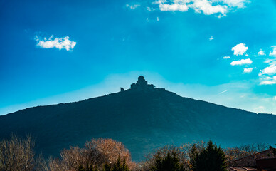 Jvari Monastery appears in dark silhouette atop a steep hill, rising above sunlit trees and set...
