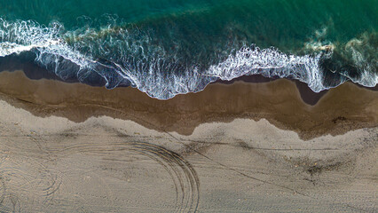 Aerial view of a beach with waves crashing onto the shore and tire tracks in the sand