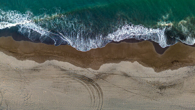 Aerial view of a beach with waves crashing onto the shore and tire tracks in the sand - Powered by Adobe