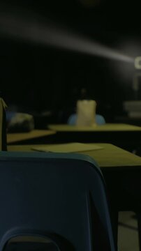 empty classroom desk with projector beam, dim light slicing through dark room, candle glowing on center desk, scattered papers and blue chairs, silent study