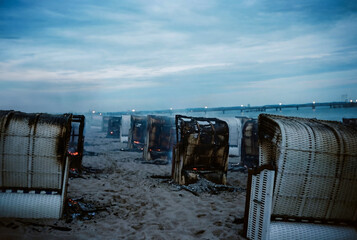 Moody photograph of burned beach chairs standing in rows on a deserted shoreline at dusk. Charred wicker structures, glowing embers, and drifting smoke create a dramatic, cinematic atmosphere.
