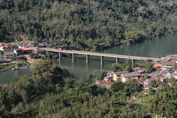 Panoramic autumn mountain landscape featuring a high peak view over a misty forest valley with green trees and a lake under a cloudy sky in Laos