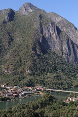 Panoramic autumn mountain landscape featuring a high peak view over a misty forest valley with green trees and a lake under a cloudy sky in Laos