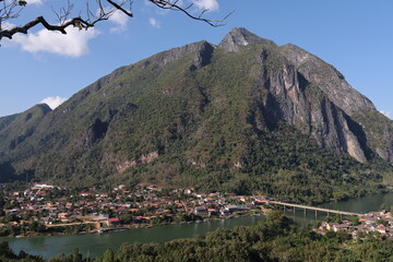 Panoramic autumn mountain landscape featuring a high peak view over a misty forest valley with green trees and a lake under a cloudy sky in Laos