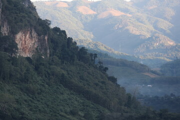 Panoramic autumn mountain landscape featuring a high peak view over a misty forest valley with green trees and a lake under a cloudy sky in Laos