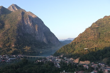 Panoramic autumn mountain landscape featuring a high peak view over a misty forest valley with green trees and a lake under a cloudy sky in Laos