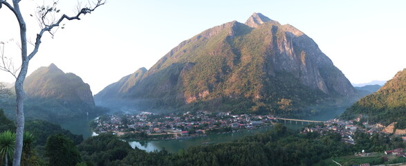 Panoramic autumn mountain landscape featuring a high peak view over a misty forest valley with green trees and a lake under a cloudy sky in Laos