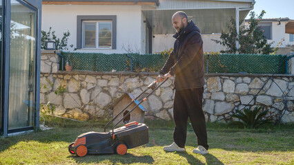 A middle-aged man mowing the grass in his garden with a lawnmower on a winter day