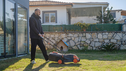 A middle-aged man mowing the grass in his garden with a lawnmower on a winter day