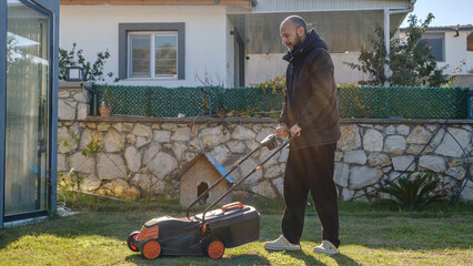 A middle-aged man mowing the grass in his garden with a lawnmower on a winter day