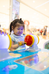 A cute little Asian girl is playing a coin-operated game machine in a shopping mall,Cute little girl adjusting knob and pressing red button while looking at screen of game machine at leisure center