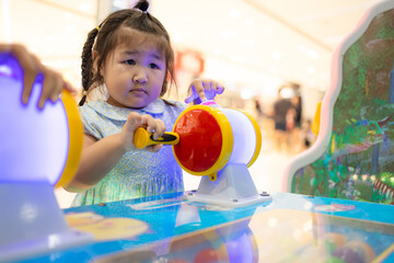 A cute little Asian girl is playing a coin-operated game machine in a shopping mall,Cute little girl adjusting knob and pressing red button while looking at screen of game machine at leisure center