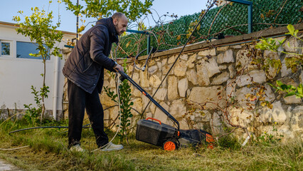 A middle-aged man mowing the grass in his garden with a lawnmower on a winter day