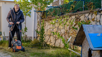A middle-aged man mowing the grass in his garden with a lawnmower on a winter day
