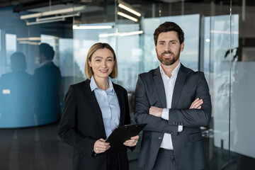 Two smiling business professionals, a woman holding a clipboard and a man with crossed arms, standing confidently in a modern glass office portraying teamwork and success