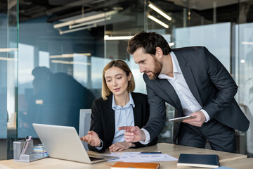 Businesswoman and businessman discussing project on a laptop, working together and analyzing data in a modern office, showing teamwork and professional collaboration
