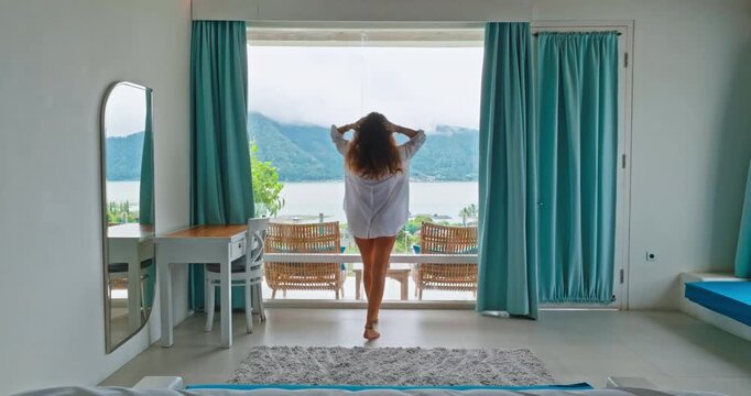 Beautiful young woman in white shirt walking bright hotel room, relax and enjoy serene morning landscape lake view from a wide window, relaxation, travel, and vacation lifestyle in Bali. Back view