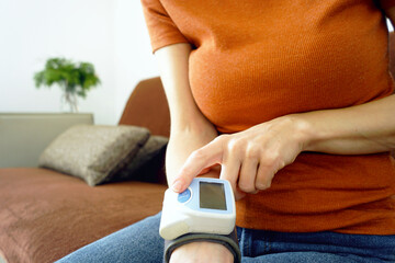 Young woman checking a digital blood pressure reading on a wrist device while seated comfortably indoors. Represents remote patient monitoring, vital signs tracking, and empowered personal healthcare.