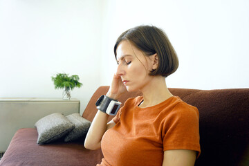 Woman touching her temple with a hand wearing a blood pressure monitor, experiencing a headache symptoms. Illustrates hypertension-related discomfort, migraine triggers, and vascular tension.