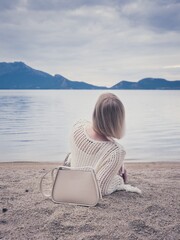 Back view of a woman sitting on a sandy beach, featuring a stylish beige handbag and a mountain lake view.