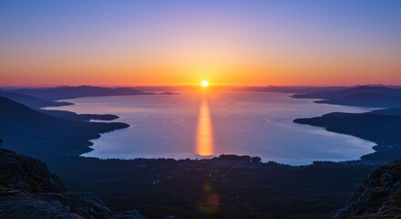 Brilliant sun descends over expansive water body framed by silhouetted mountains and foreground rocks