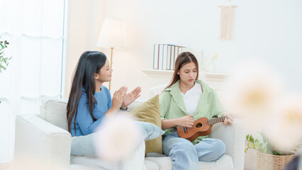 Two young Asian women sharing a joyful moment at home. One girl is playing the ukulele while her friend claps along and cheers. They are sitting on a cozy sofa in a bright, minimalist living room.