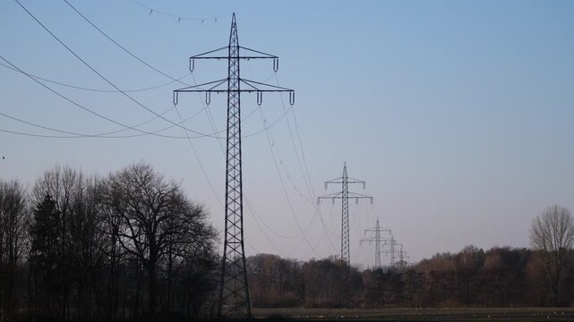 Several power poles or pylons with power lines standing in a row, slightly orange-blue evening sky