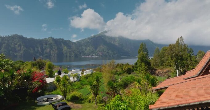 Balinese style traditional villa hotel resort with red roof tiles nestled in a lush tropical garden, offering stunning panoramic views of Lake Batur and Mount Abang in background. Aerial drone flight