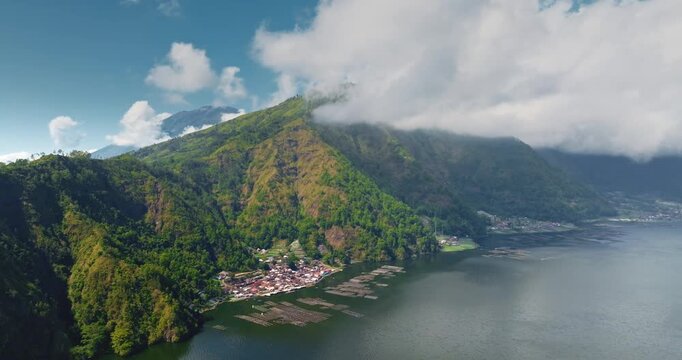 Indonesia, Bali, Abang village and traditional aquaculture rafts floating on calm water of Lake Batur, green hills and clouds covering the peaks of Mount Abang volcano. Aerial drone flight panorama