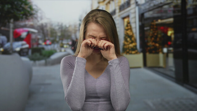 Young woman crying on a city street with christmas decorations in the background, expressing emotions during wintertime in an urban outdoor setting.