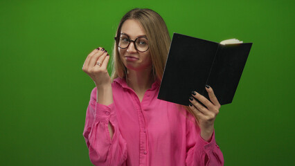 Woman in pink shirt against green background reading book and making money gesture, appearing...