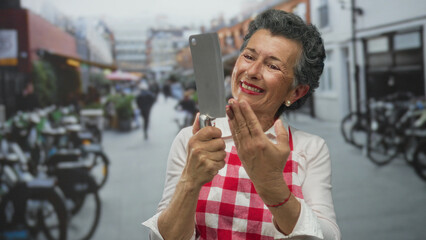 Senior woman with grey hair in apron holding a knife and smiling on an urban street, portraying cheerful culinary energy in lively city setting.