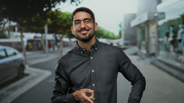 Young handsome man smiling and thinking on an urban street, showcasing a casual style with glasses and beard, set against a city background indicating a vibrant town atmosphere.